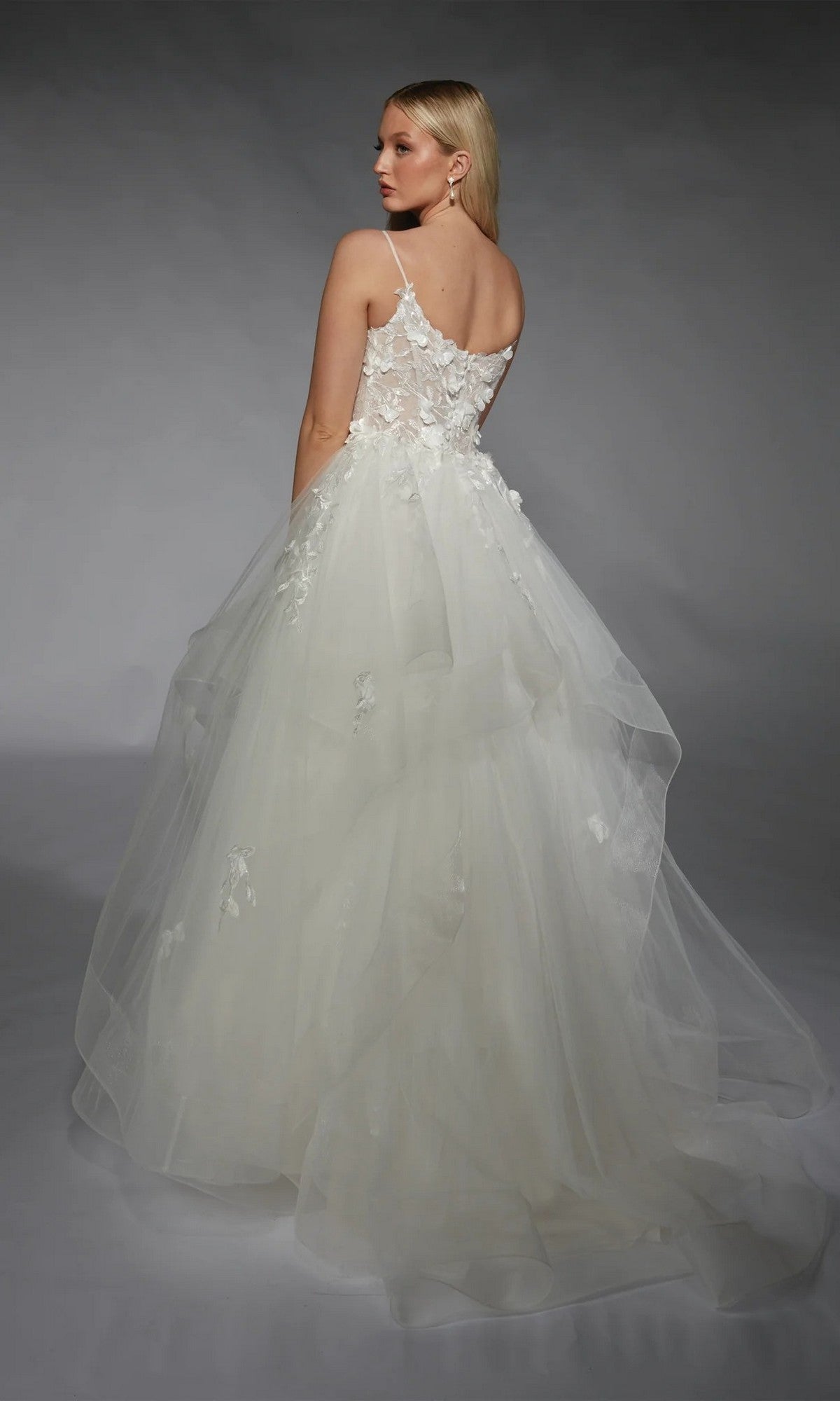 A woman stands against a gray background, facing away from the camera, wearing the Alyce 7131 long white dress—a sleeveless bridal ball gown with a tulle ruffled skirt and 3D floral lace appliques.